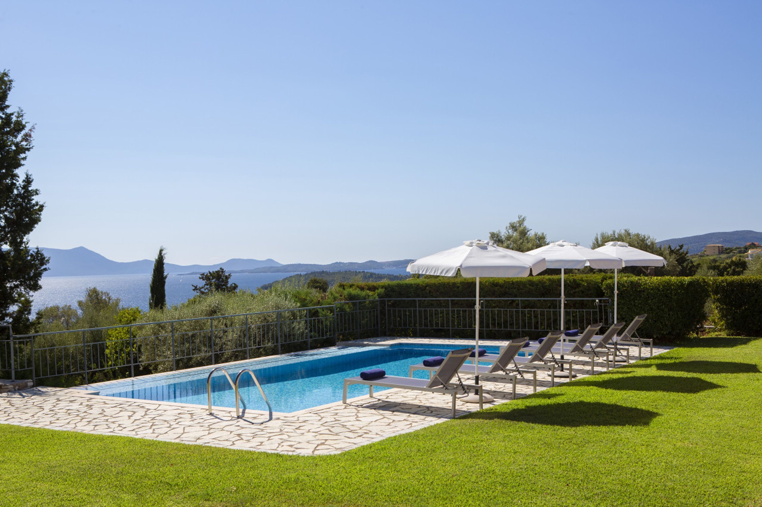 Una piscina con sillas de descanso y sombrillas, rodeada de un césped cuidado y vistas al mar y a las montañas.