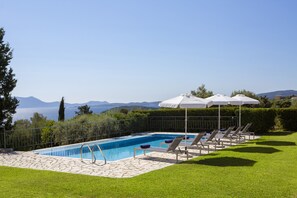 Una piscina con sillas de descanso y sombrillas, rodeada de un césped cuidado y vistas al mar y a las montañas.