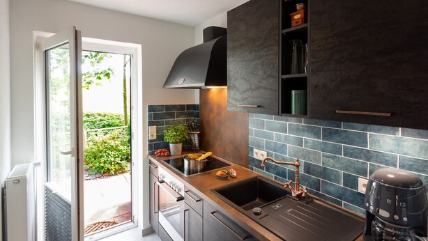 Una cocina moderna con horno empotrado, lavamanos y estufa. Se ve un ventanal con vistas a la vegetación.