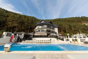 A multi-story building with a pool in front, surrounded by trees and a mountainous backdrop.