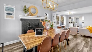 A modern dining area with a wooden table, six chairs, a black brick fireplace, and a gold chandelier.