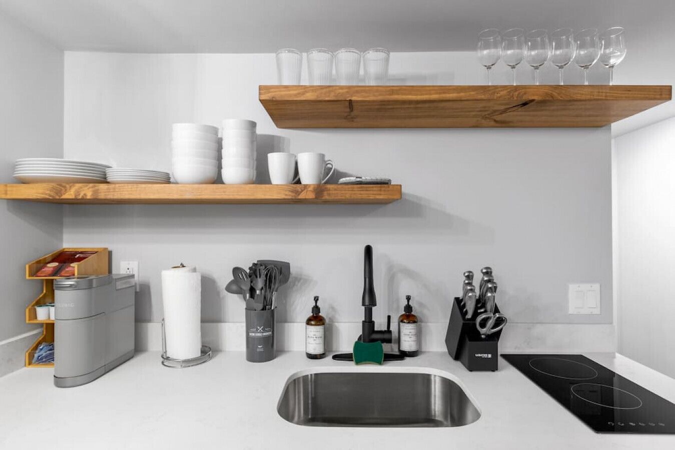 A modern kitchen with white countertops, a stainless steel sink, wooden shelves with dishes, and various kitchen utensils.