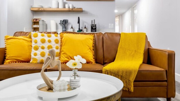 Un salon moderne avec un canapé en cuir marron, des coussins à motifs jaunes et blancs, un vase blanc contenant une fleur et une sculpture en bois.