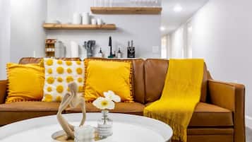 Un salon moderne avec un canapé en cuir marron, des coussins à motifs jaunes et blancs, un vase blanc contenant une fleur et une sculpture en bois.