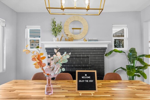 A modern dining area with a wooden table, a welcome sign, a fireplace, and decorative plants. - 4016 Main (NEW) · 5b2b Newly Remodeled Entire House close to UW & DT (Seattle)