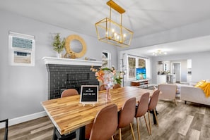 A modern living room with a fireplace, a dining area with a wooden table and chairs, and a kitchen in the background.