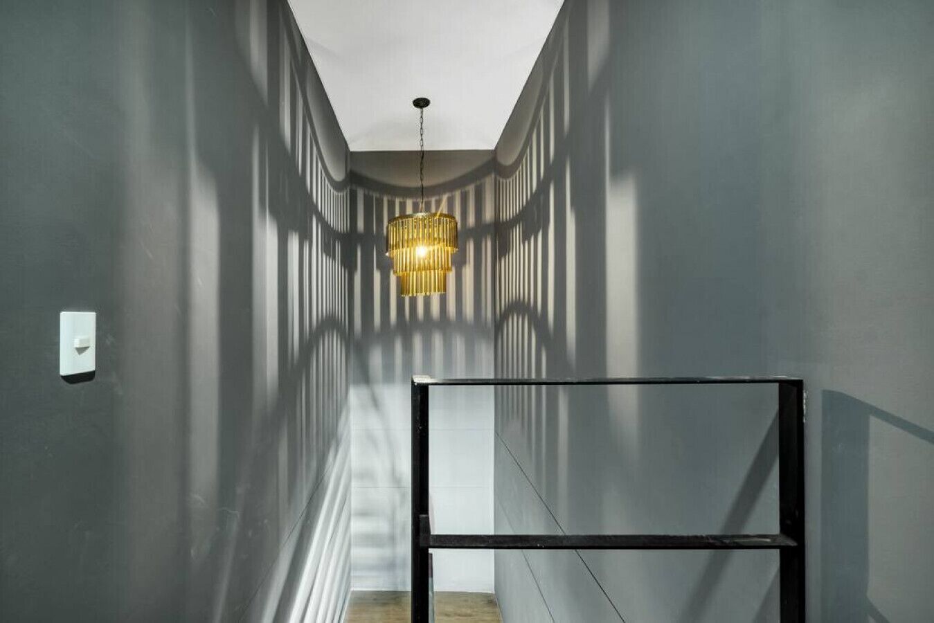 Een smalle gang met een moderne kroonluchter, een constructie van metaal en een lichtschakelaar aan de muur.