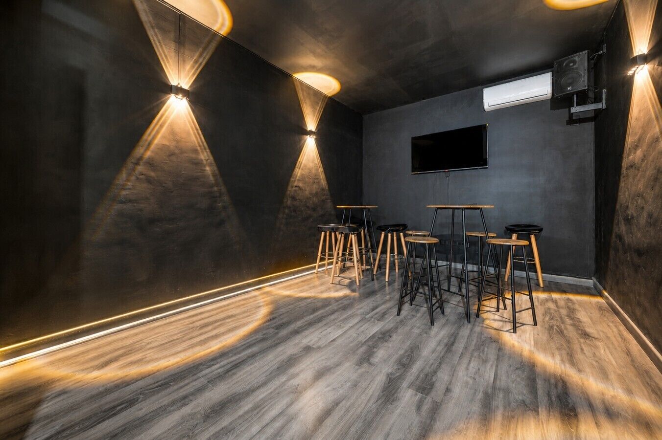 A room with a dark wall, a flat-screen TV mounted on the wall, and several wooden stools arranged around a central table.