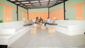 Un espacio cubierto con sofás blancos y taburetes de madera, bajo un techo de vigas de madera.