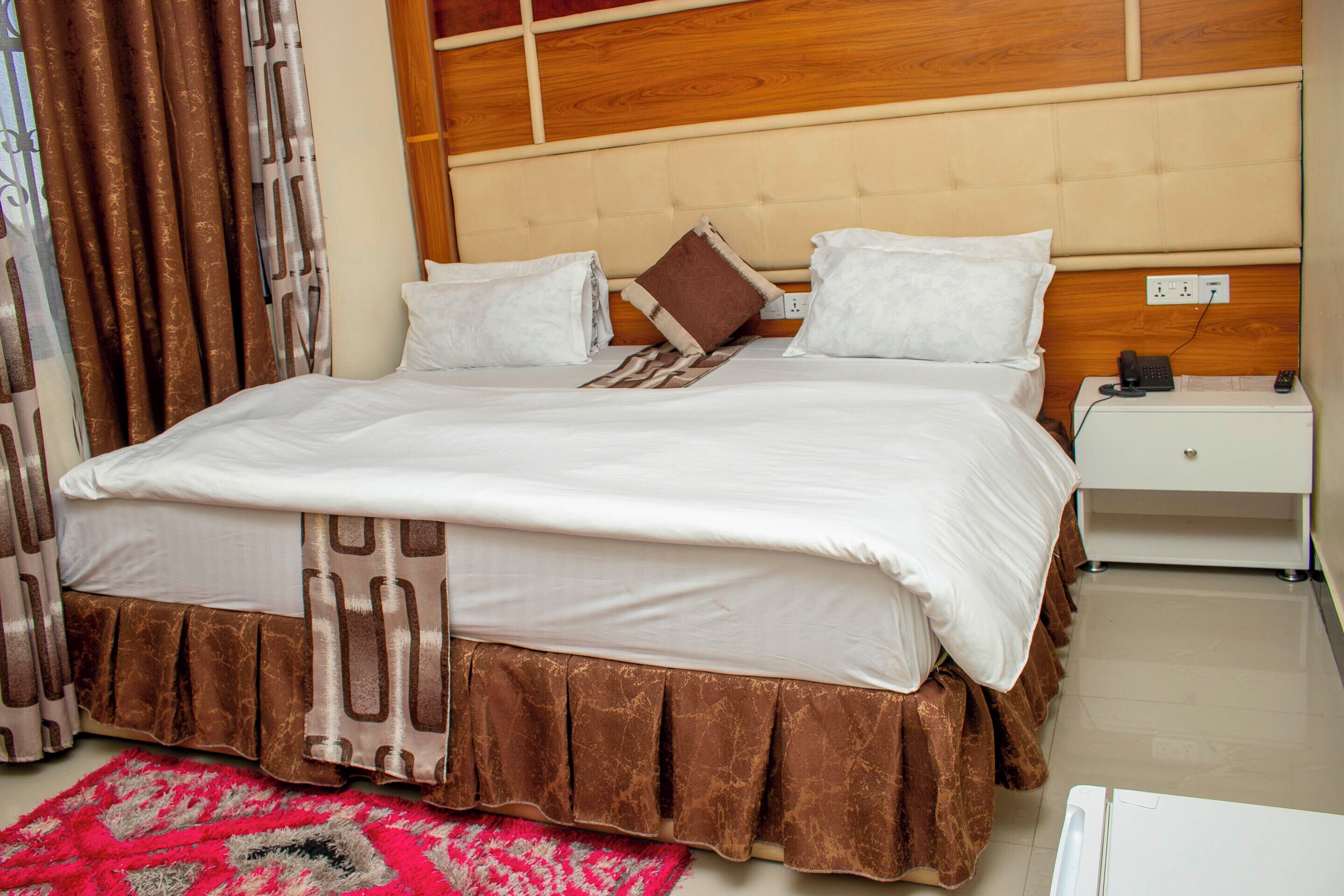 Un lit bien fait, avec du linge de lit blanc, une base de lit marron et un couvre-lit à motifs. On y trouve une table de chevet blanche avec un téléphone et une télécommande, ainsi qu’une tête de lit en bois.