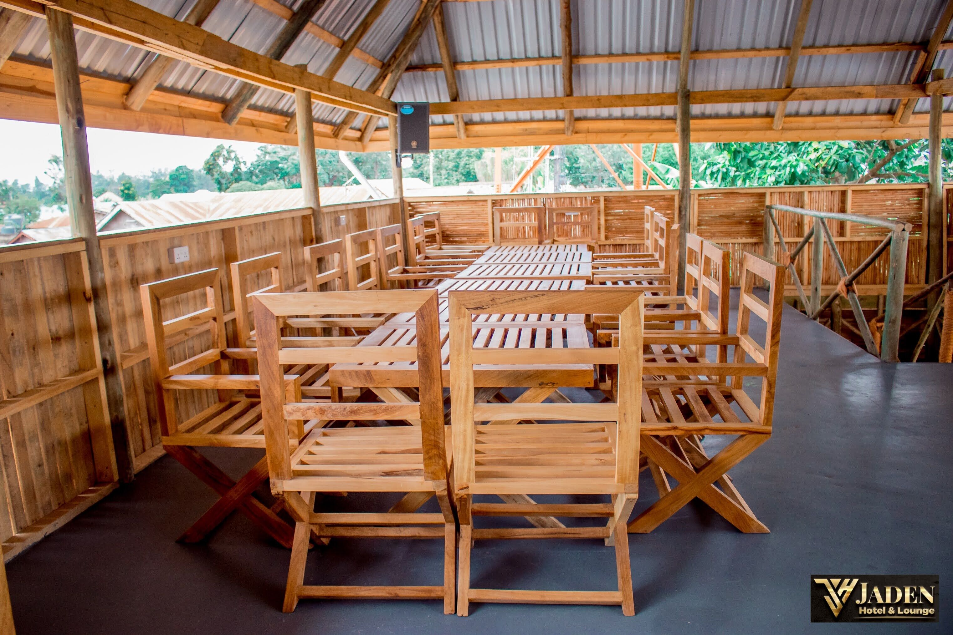 A covered outdoor seating area with wooden tables and chairs, a metal roof, and a view of greenery outside.
