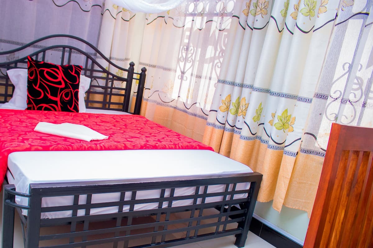 A neatly made bed with a red bedspread and white pillows, a wooden headboard, and a curtain with a floral pattern.