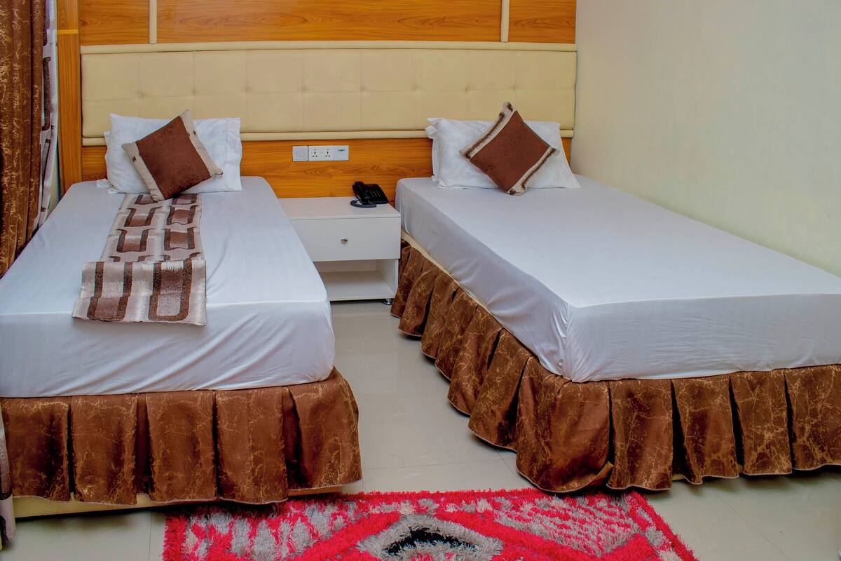 A hotel room with two beds, each with a brown bed skirt and white linens. There is a nightstand between the beds with a phone and a lamp.