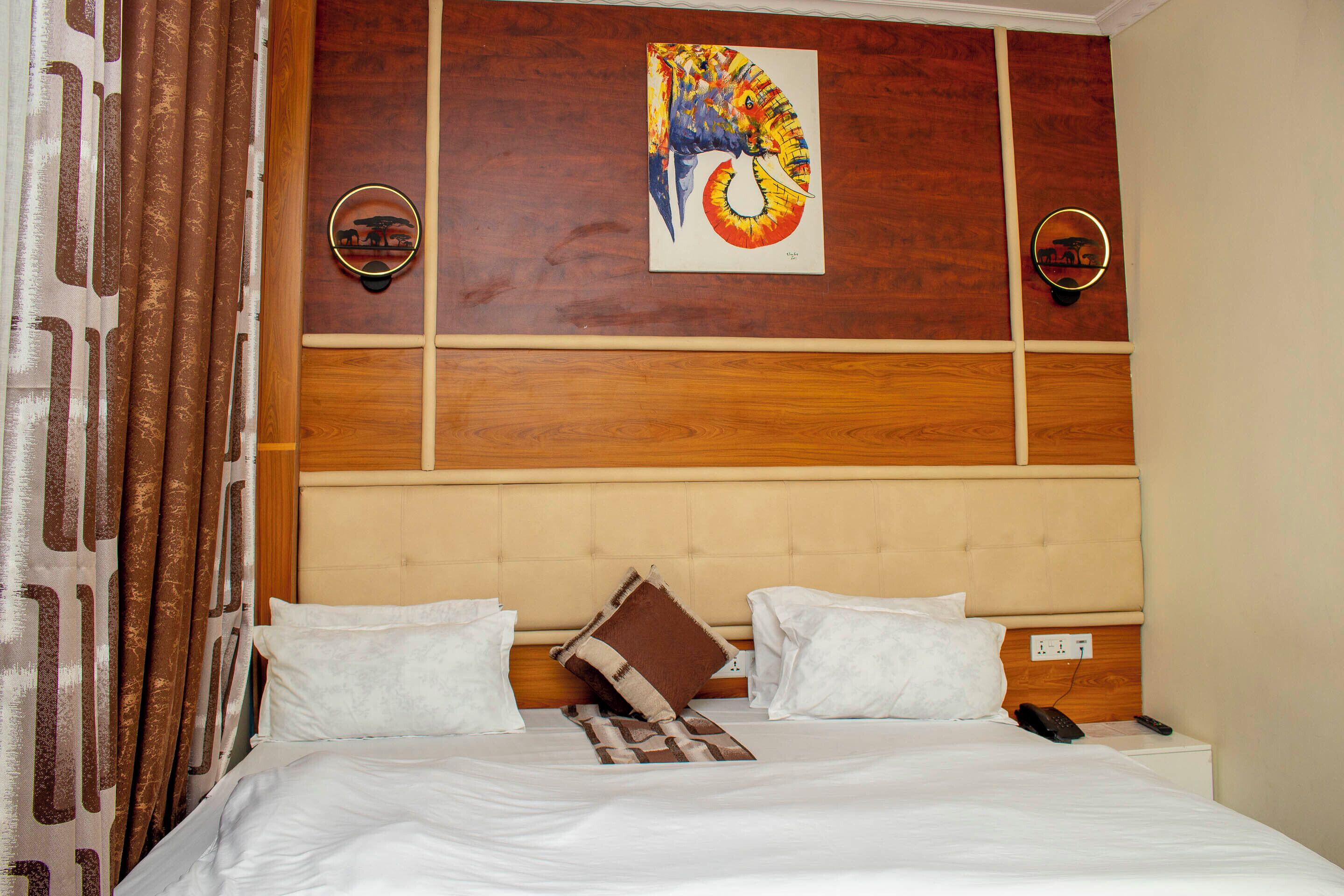 A hotel room with a bed, white bedding, a headboard with a wooden panel design, a painting above the bed, and two wall-mounted phones.