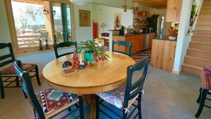 A dining area with a wooden table and chairs, a hanging light fixture, and a view of the kitchen and living area.