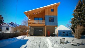 A two-story wooden house with a balcony, a garage, and a snow-covered driveway.