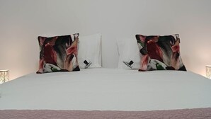 A bed with a white duvet, two decorative pillows with a floral pattern, and a painting of a violinist on the wall above. - Casa Típica no Centro do Porto (PORTO)