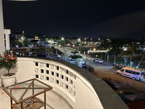 Un balcón con una mesa de vidrio y una silla de mimbre, con vista a una calle concurrida por la noche, con edificios iluminados y banderas.