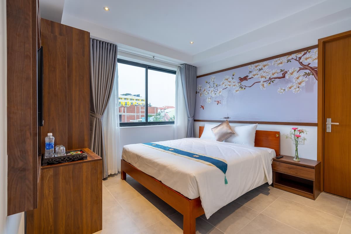 A hotel room with a large bed, wooden wardrobe, a nightstand, a bedside table, and a window with curtains.