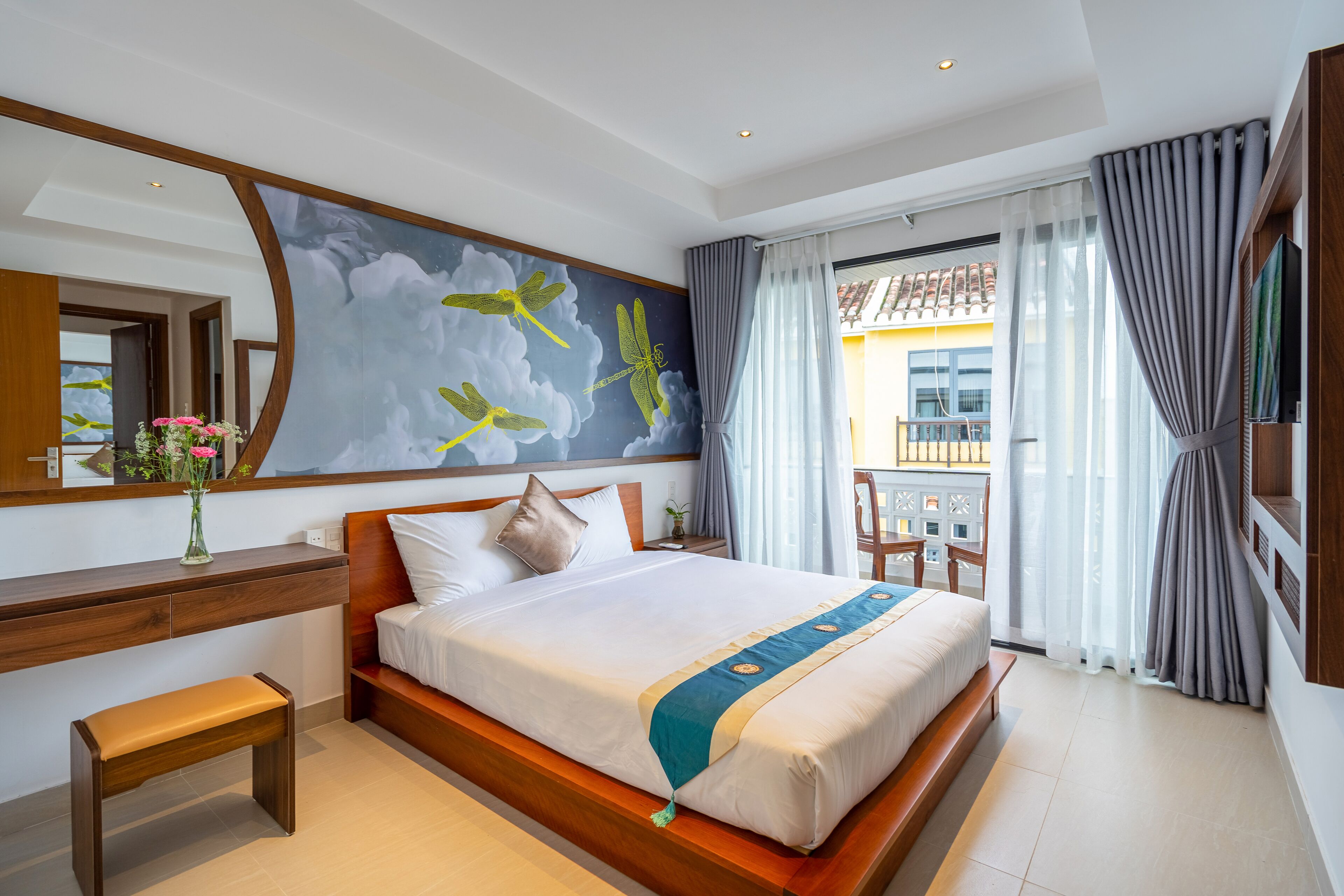 Habitación de hotel con una cama grande, un escritorio de madera, un banco, un espejo amplio y vistas al balcón exterior.