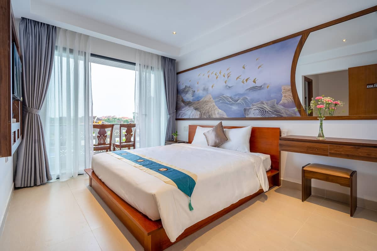 A hotel room with a large bed, a wooden desk, a chair, a painting on the wall, and a view through the window.