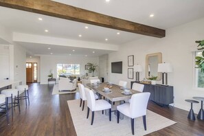 A spacious dining area with a long table, white chairs, and a wooden ceiling beam.