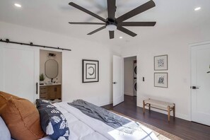 Un dormitorio con cama, ventilador de techo, banco y puerta que da a un área de lavandería.