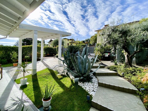 A covered patio area with a white pergola, a stone pathway, and a well-maintained lawn. - Villa Chiara (Polpenazze del Garda)