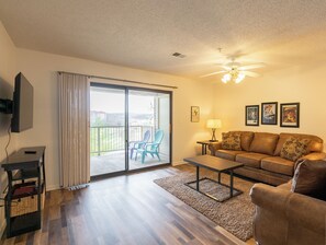 A living room with a brown leather couch, a coffee table, a TV on a stand, and a sliding glass door leading to a balcony with chairs.