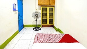 Una habitación con puerta azul, un armario de madera, un ventilador de pie y una cama con colcha estampada.