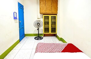 Une pièce avec une porte bleue, une armoire en bois, un ventilateur sur pied et un lit recouvert d’une couvre-lit à motifs.
