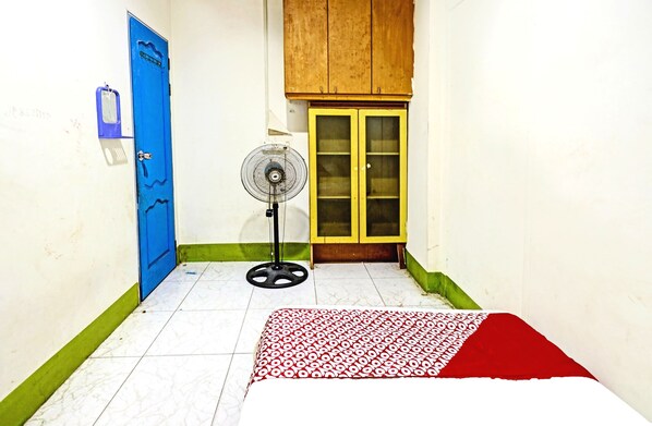 A room with a blue door, a wooden cabinet, a standing fan, and a bed with a patterned bedspread.