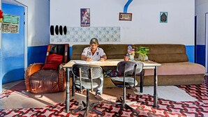 A room with a brown sofa, a table, and two chairs. There is a blue door on the left and a wall with framed pictures and a decorative panel. - Spot On 91500 Suci Homestay Syariah (Batam)