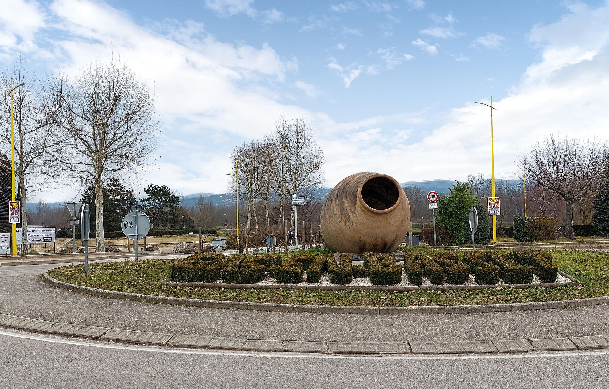Een rotonde met een grote, cilindrische sculptuur in het midden, omringd door vorm gesnoeid heggen en bomen.