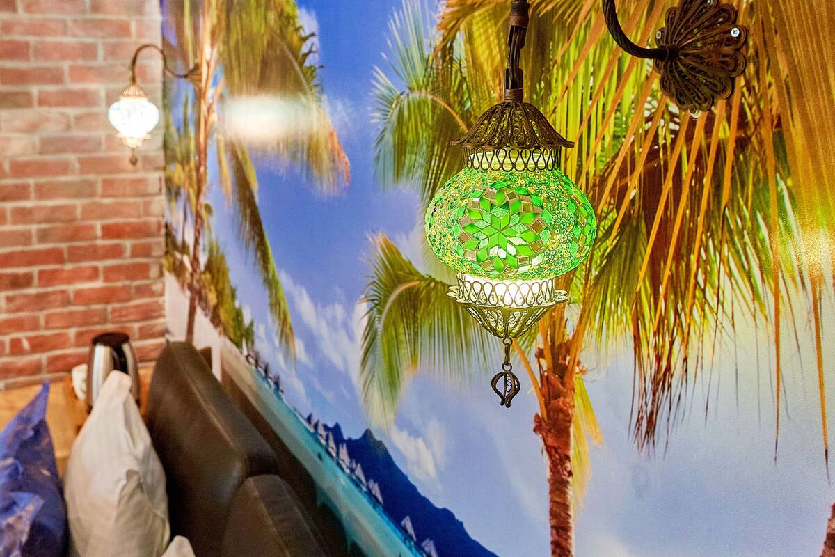 A room with a mural of palm trees and mountains, a brick wall, a black leather sofa, and a hanging green lantern.