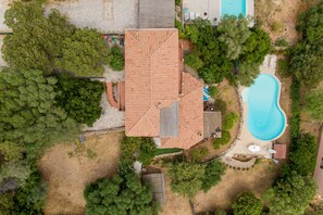 An aerial view of a property with a swimming pool, a large house with a tiled roof, and surrounding greenery.
