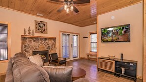Una sala de estar acogedora con chimenea de piedra, un sofá grande de cuero, un ventilador de techo de madera y un televisor sobre un mueble de madera.