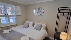 Quarto com cama, mesa de cabeceira com abajur, espelho na parede e janela com persianas.