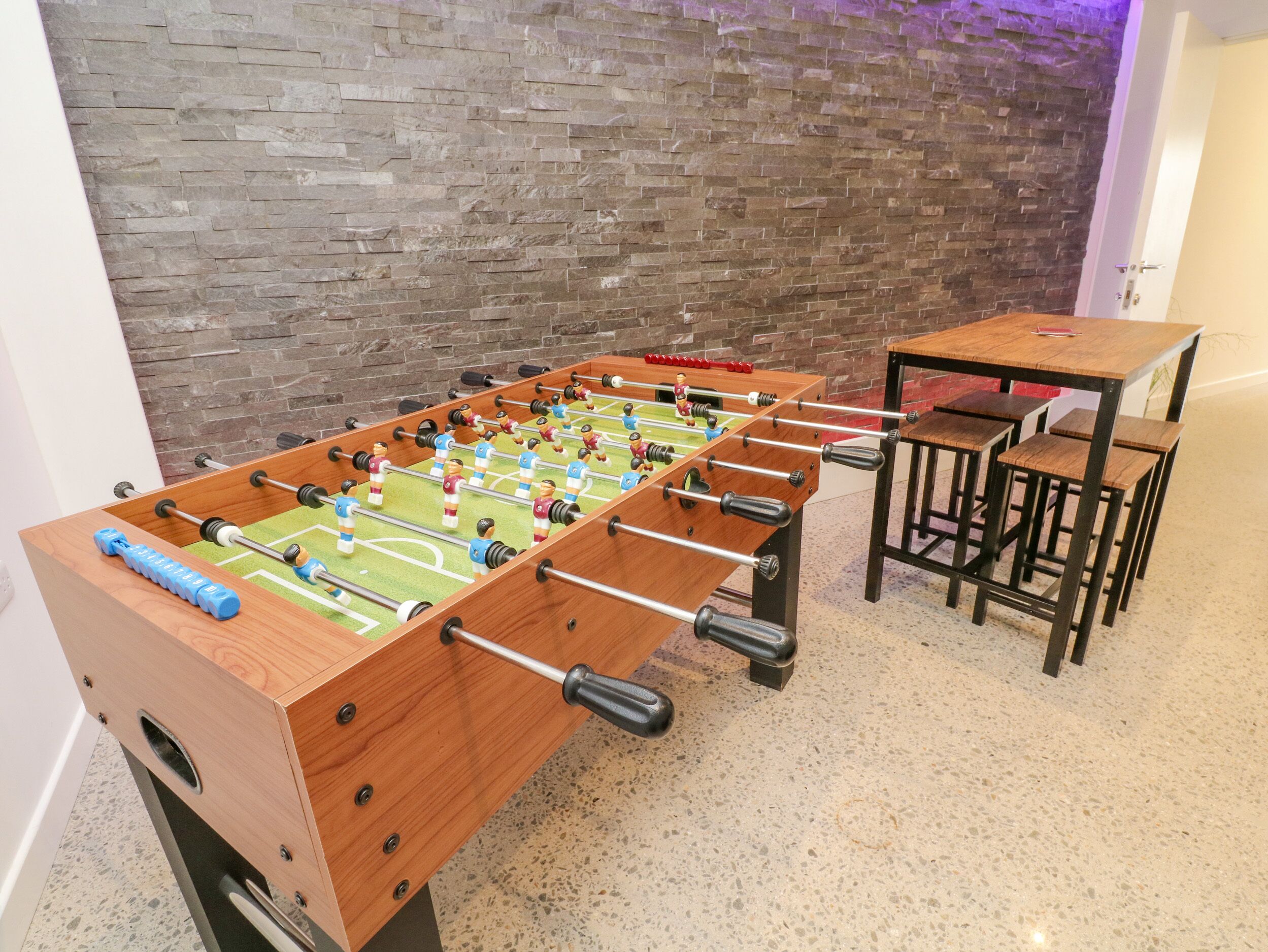 A foosball table with a brick wall backdrop and a small wooden table with chairs beside it.