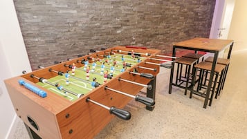 A foosball table with a brick wall backdrop and a small wooden table with chairs beside it.