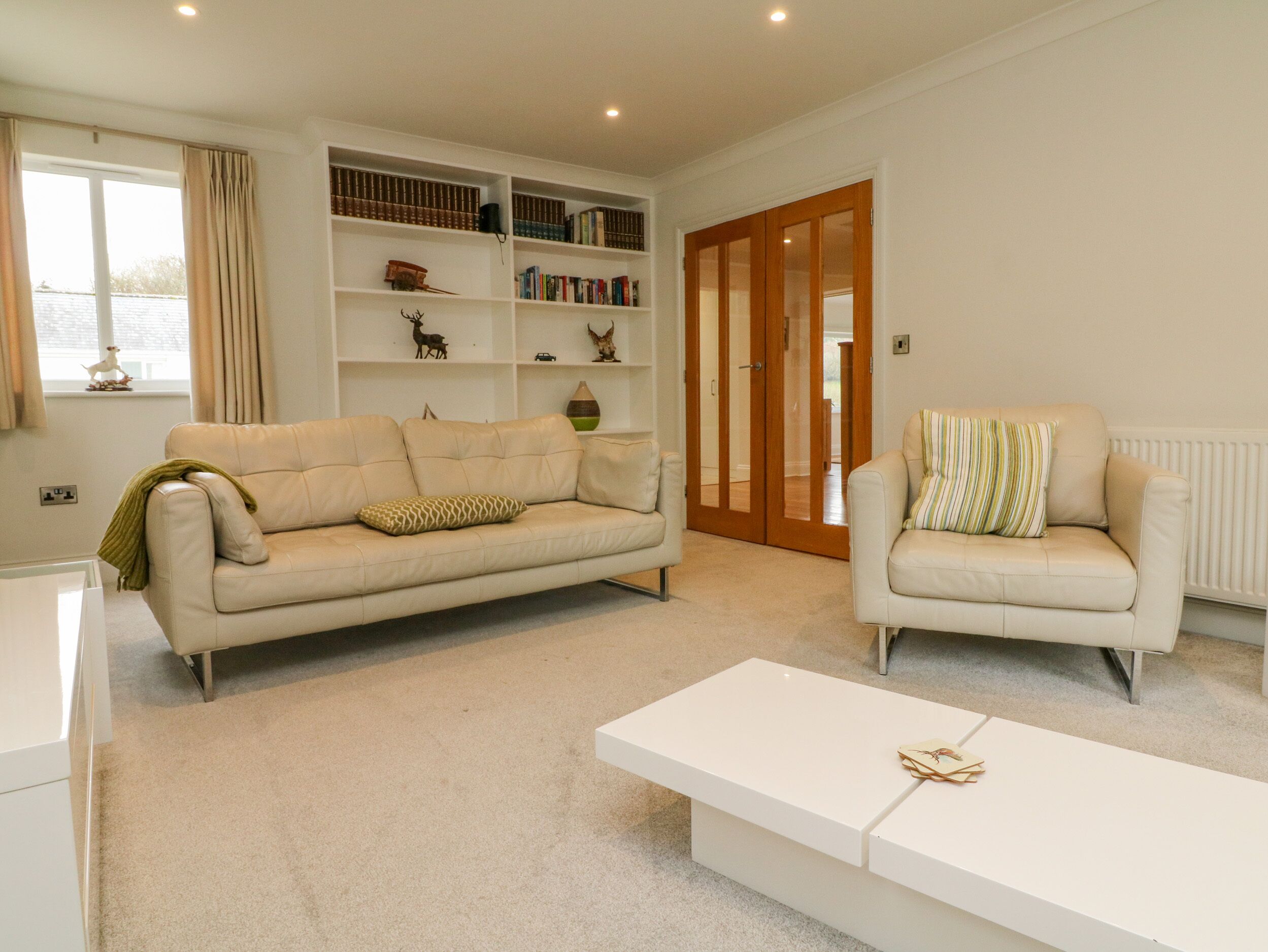 A living room with a beige sofa, a white armchair, a white coffee table, and a bookshelf filled with books and decorative items.