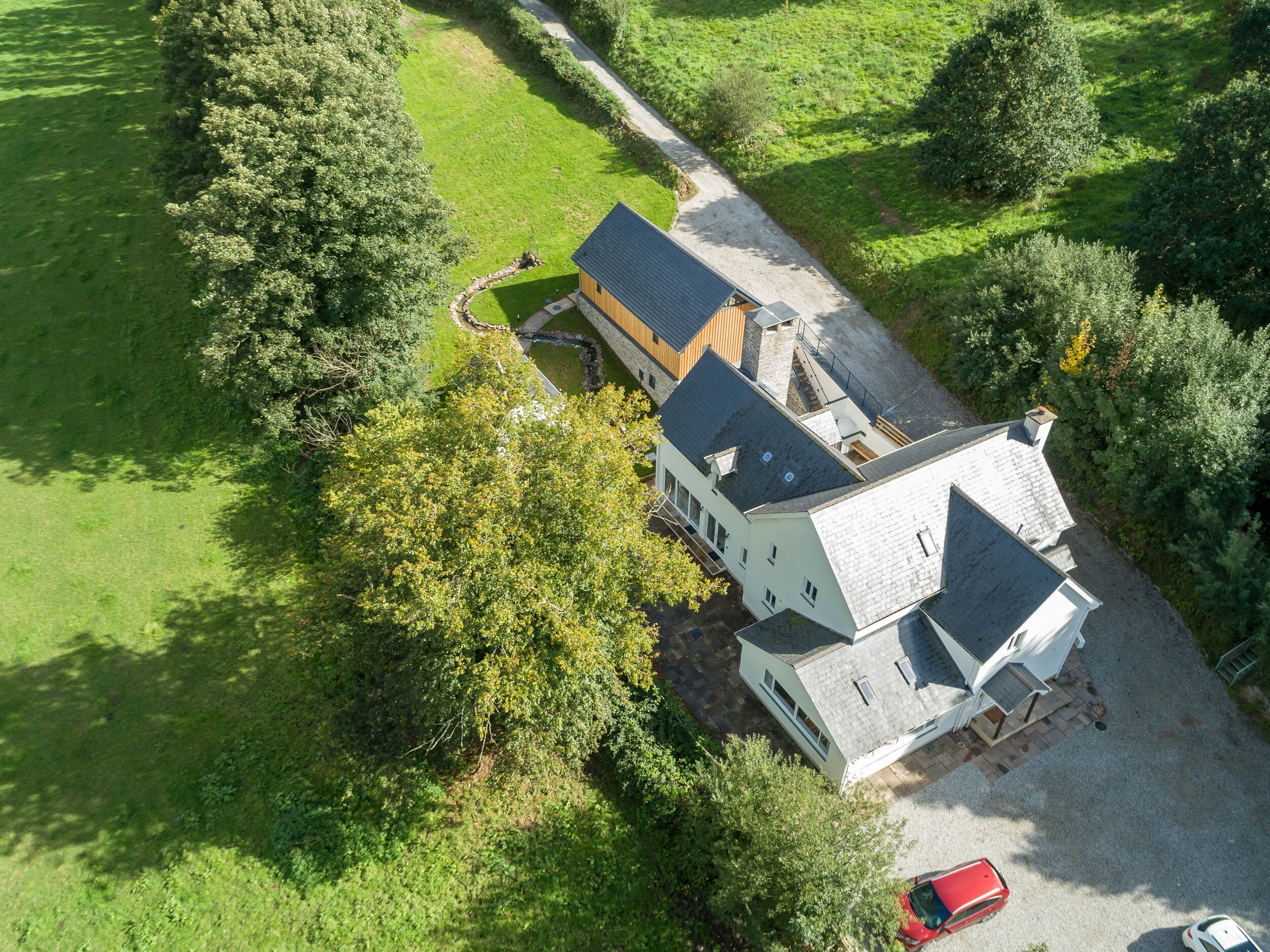 A large, two-story house with a sloped roof, surrounded by lush greenery and a gravel driveway.
