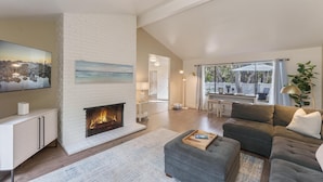 A modern living room with a fireplace, a large flat-screen TV, a grey sectional sofa, and a view of the outdoors through a large window. - 3880 Casa Azul - Near Hiking Trails, Golf & Town (Pebble Beach)