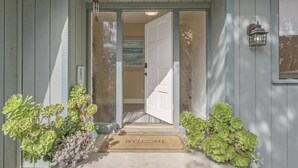 A wooden house entrance with a welcome mat, a door, and a small garden with green plants. - 3880 Casa Azul - Near Hiking Trails, Golf & Town (Pebble Beach)