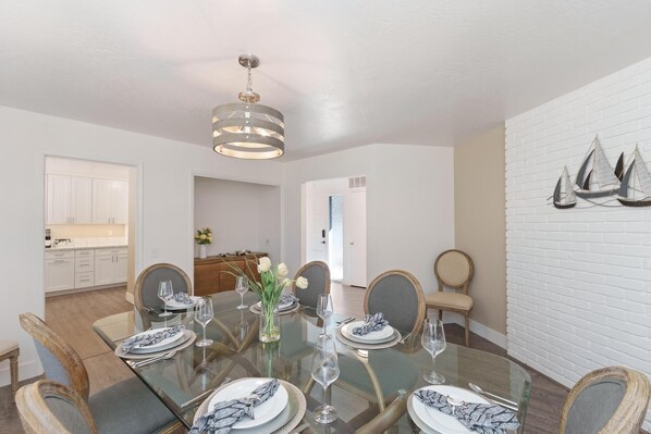 A dining area with a glass table set for six, surrounded by wooden chairs. A chandelier hangs from the ceiling, and a white brick wall with sailboat decorations is visible. - 3880 Casa Azul - Near Hiking Trails, Golf & Town (Pebble Beach)