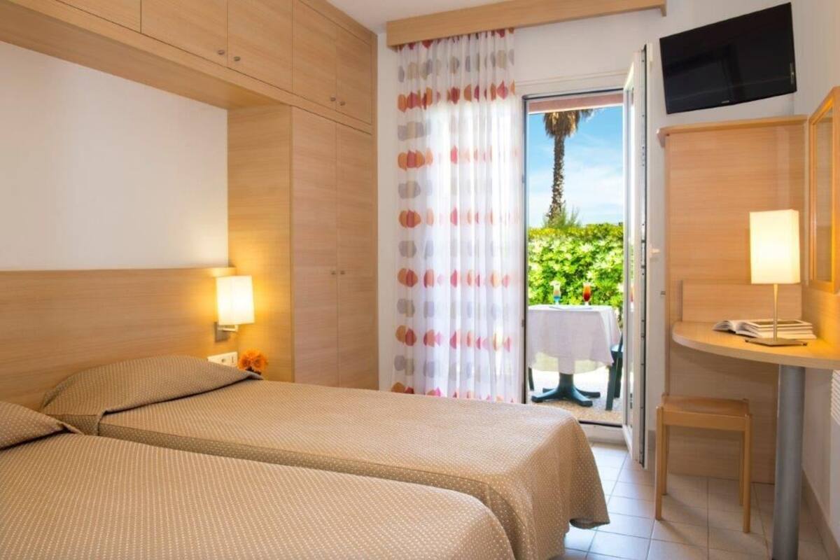 Quarto de hotel com duas camas, televisão, escrivaninha com luminária e varanda com mesa e cadeiras.