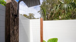 Una ducha exterior moderna con un poste de madera y paredes de azulejos blancos, rodeada de abundante vegetación.