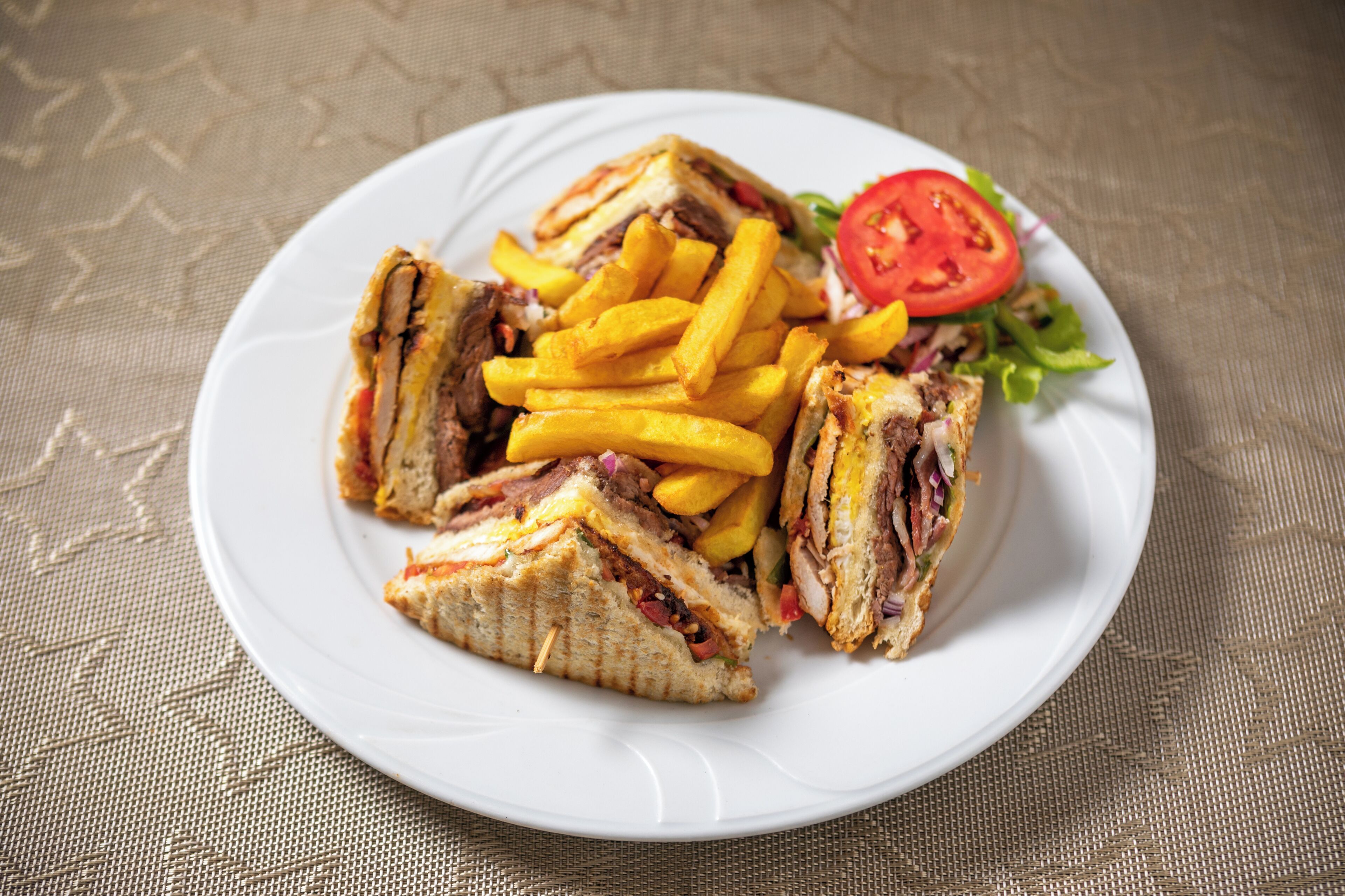 A plate with a sandwich, fries, and a slice of tomato on a textured tablecloth.
