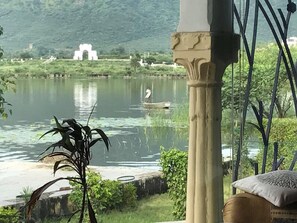 A serene riverside view with a white arch structure in the background, a person rowing a boat, and a column with a cushion. - Hotel Vishalas The Raj Kesar Regency (Udaipur)