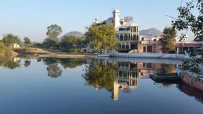 A hotel with a prominent tower and arched windows, situated by a calm body of water with clear reflections. - Hotel Vishalas The Raj Kesar Regency (Udaipur)
